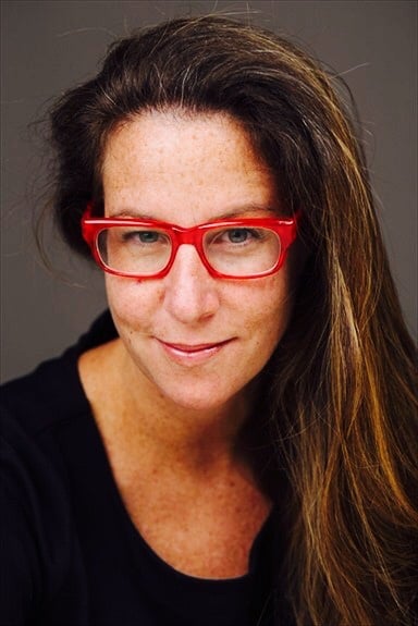 Headshot of Hillary Black: A light-skinned, freckled woman with long brunette hair wearing red glasses and a black shirt. She is smiling softly.