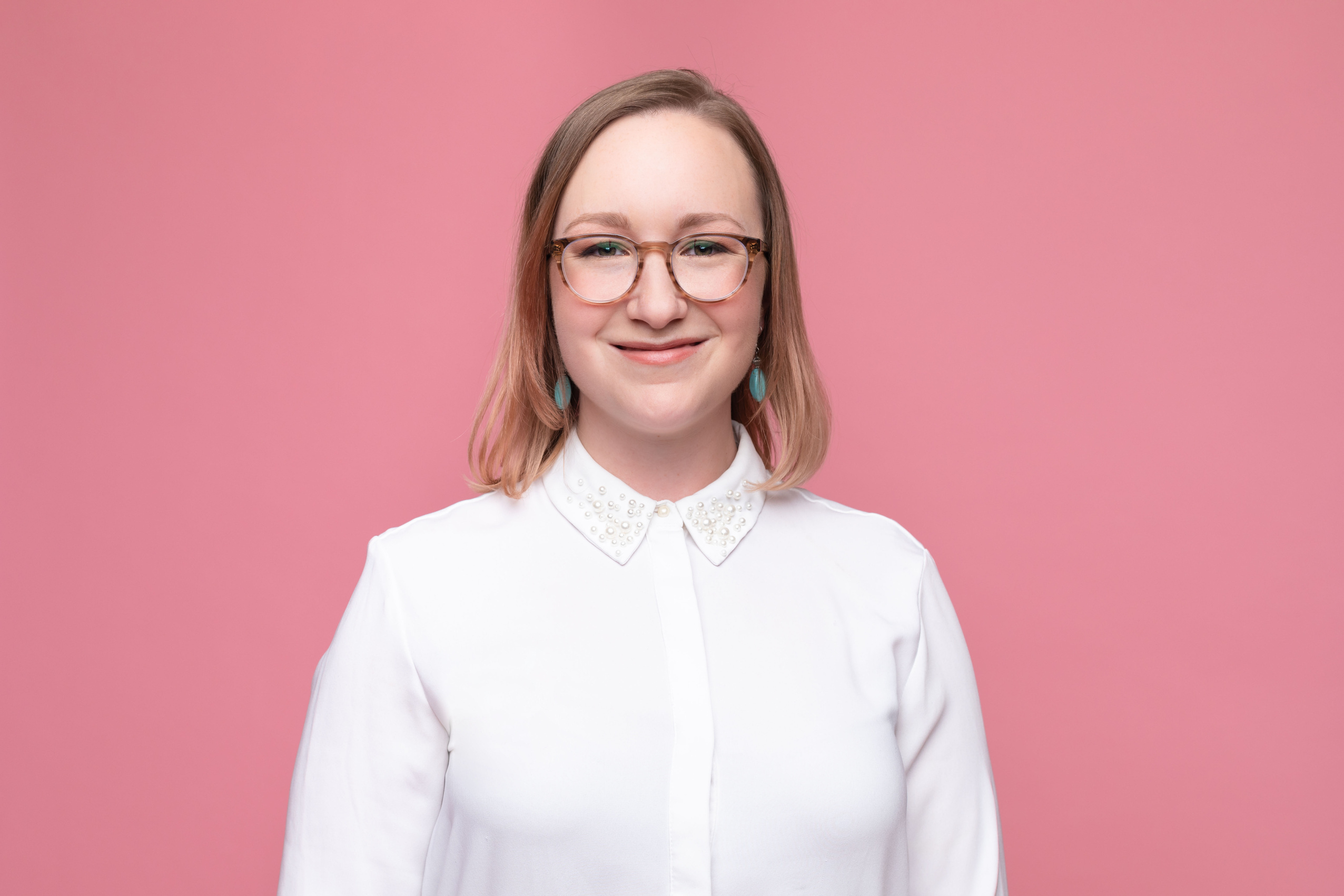 Headshot of Kathryn Freund: A light-skinned woman with brown glasses and short blonde hair wearing a white button-up shirt in front of a pink background. She is smiling softly.